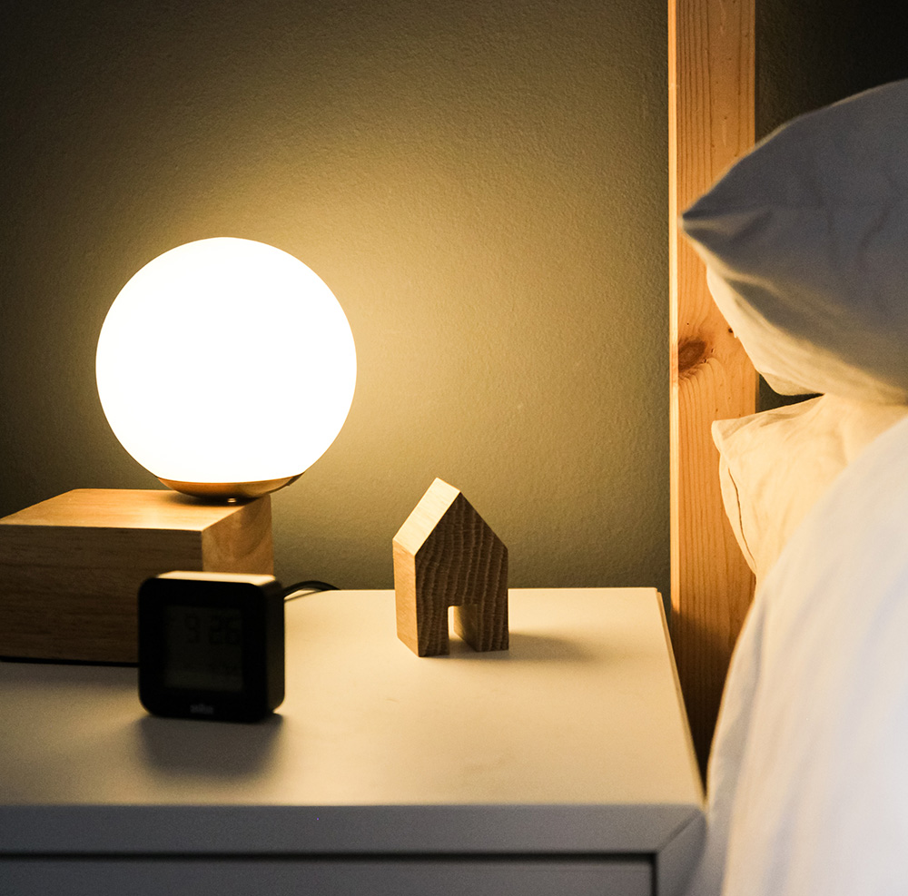 detail of a bedside table next to a bed with a lamp digital clock and wooden house