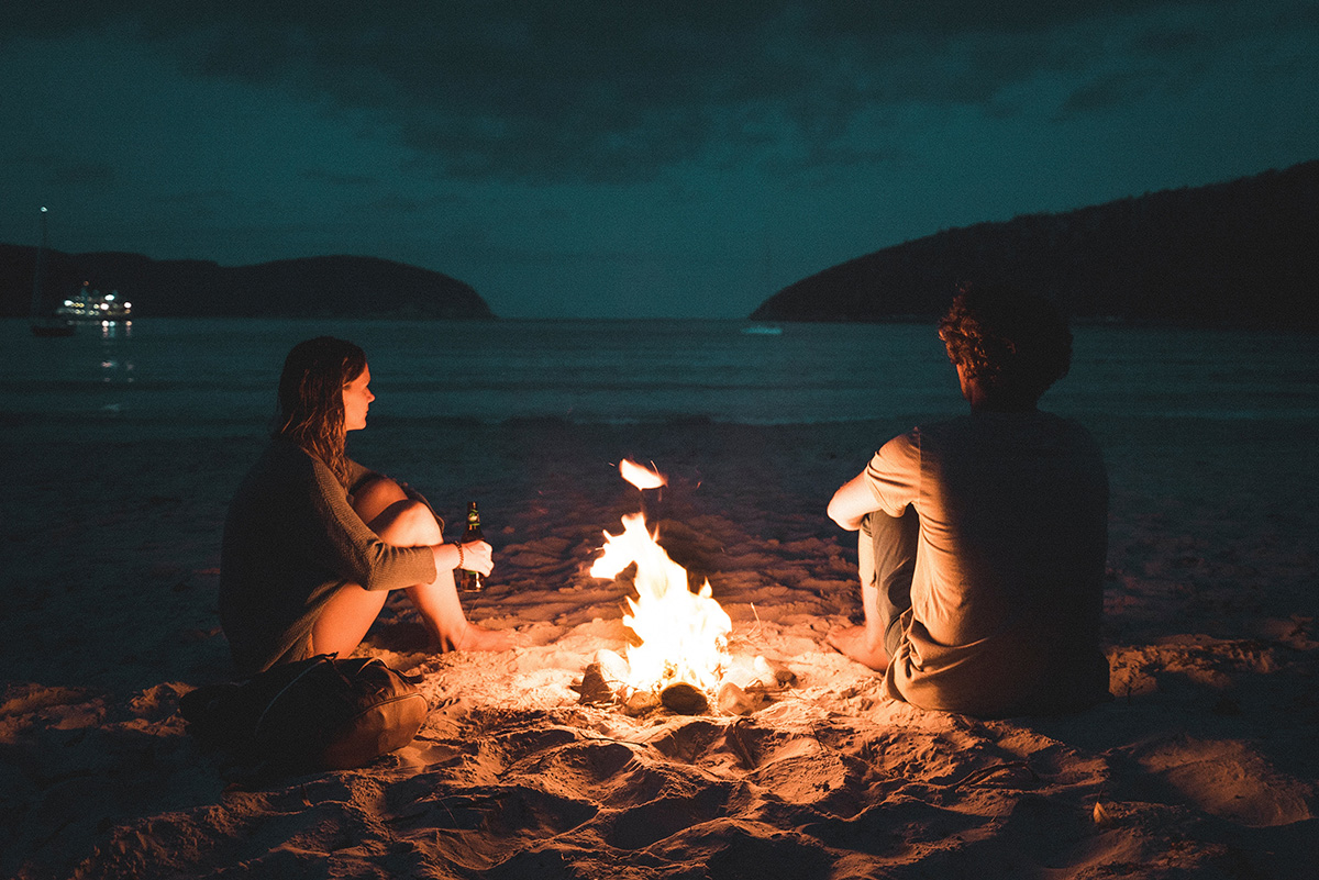 two people sitting in the beach at night around a fire sharing a romantic moment